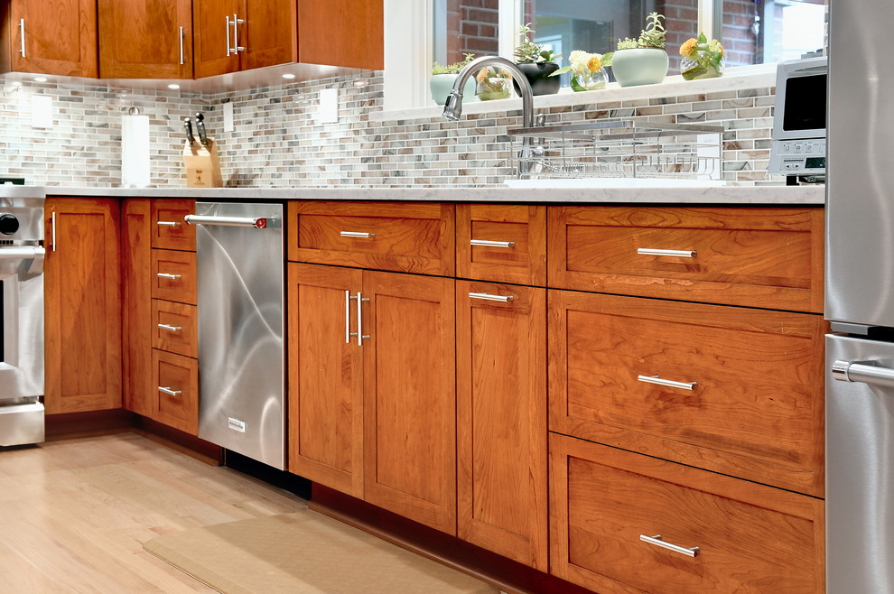 This is an example of a mid-sized transitional l-shaped separate kitchen in DC Metro with an undermount sink, glass-front cabinets, medium wood cabinets, marble benchtops, multi-coloured splashback, glass tile splashback, stainless steel appliances, light hardwood floors, no island, beige floor and white benchtop.