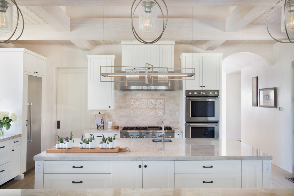 This is an example of a large mediterranean l-shaped open plan kitchen in Santa Barbara with with island, beige splashback, beige benchtop, an undermount sink, shaker cabinets, white cabinets, stainless steel appliances, stone tile splashback, light hardwood floors, beige floor and quartzite benchtops.
