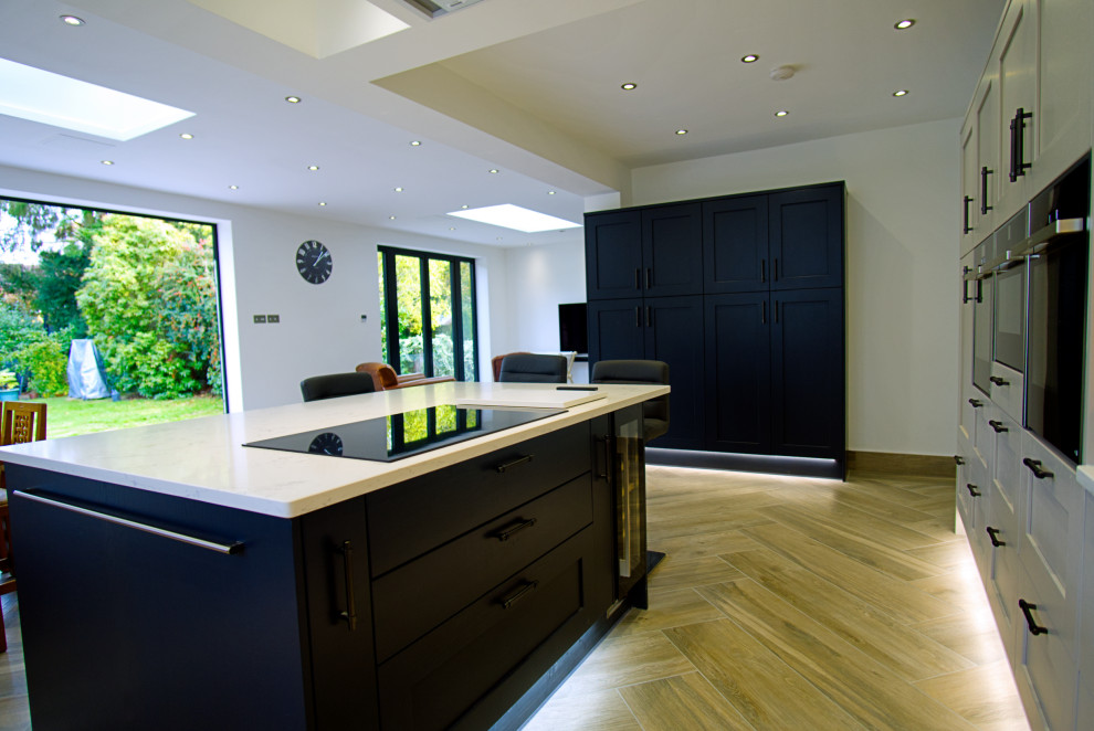 Large modern grey and black l-shaped open plan kitchen in Hertfordshire with beaded cabinets, marble worktops, white splashback, black appliances, porcelain flooring, an island, grey floors, beige worktops, feature lighting, an integrated sink and black cabinets.