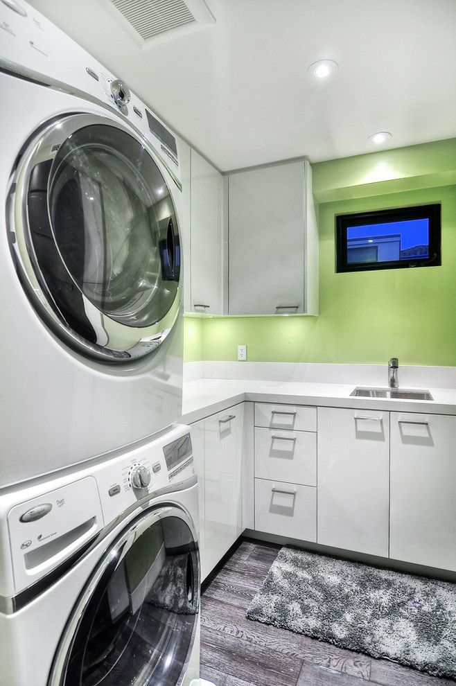Photo of a mid-sized contemporary l-shaped utility room in Orange County with green walls, white cabinets, a drop-in sink, solid surface benchtops, painted wood floors, a stacked washer and dryer and flat-panel cabinets.