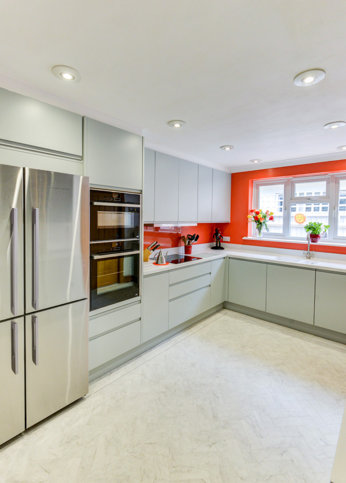 Photo of a medium sized coastal grey and white l-shaped enclosed kitchen in Sussex with a built-in sink, flat-panel cabinets, green cabinets, composite countertops, orange splashback, glass sheet splashback, black appliances, lino flooring, a breakfast bar, white floors, white worktops and feature lighting.
