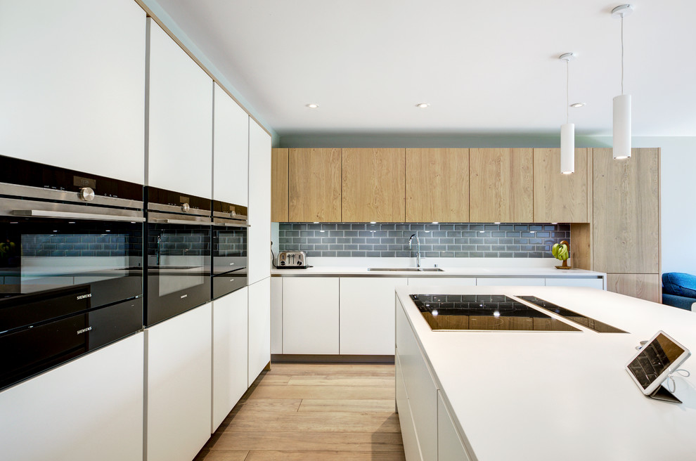 Inspiration for a mid-sized contemporary l-shaped open plan kitchen in Berkshire with flat-panel cabinets, white cabinets, solid surface benchtops, grey splashback, subway tile splashback, light hardwood floors, with island, black appliances, brown floor and a double-bowl sink.