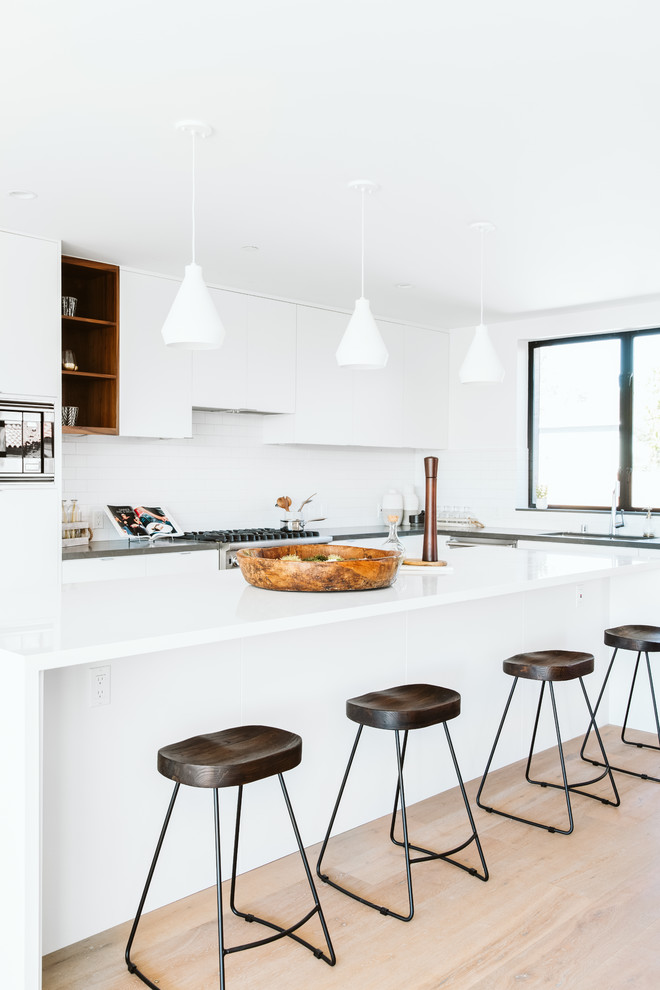 Beach style l-shaped kitchen in Los Angeles with an undermount sink, flat-panel cabinets, white cabinets, solid surface benchtops, white splashback, ceramic splashback, with island, white benchtop, light hardwood floors and beige floor.