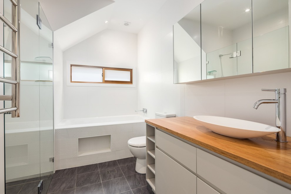 Photo of a mid-sized contemporary master bathroom in Sydney with furniture-like cabinets, white cabinets, an alcove tub, an alcove shower, a two-piece toilet, white tile, cement tile, white walls, ceramic floors, a vessel sink and wood benchtops.