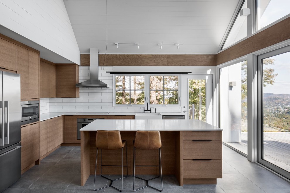 This is an example of a contemporary l-shaped kitchen in Los Angeles with a farmhouse sink, flat-panel cabinets, medium wood cabinets, white splashback, subway tile splashback, stainless steel appliances, with island, grey floor and white benchtop.