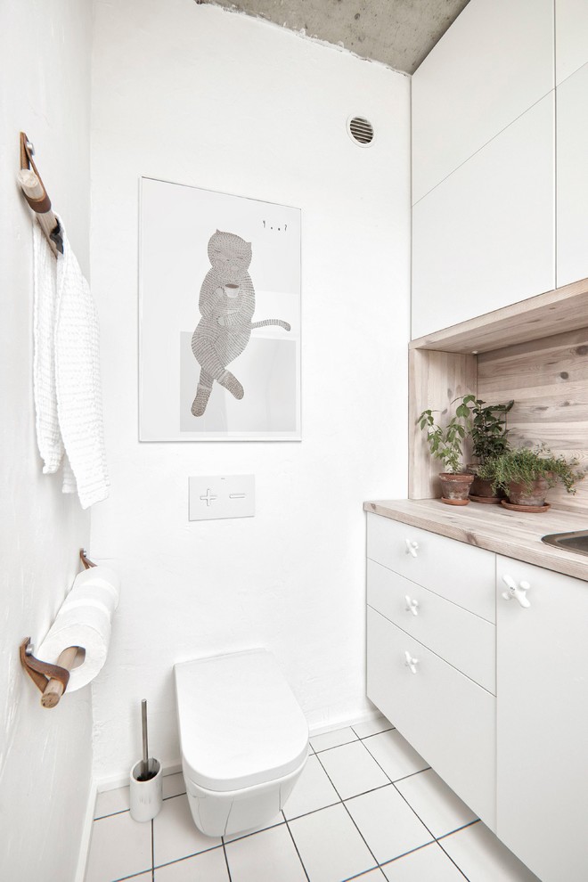 Small industrial powder room in Saint Petersburg with flat-panel cabinets, white cabinets, white walls, ceramic floors, white floor, a wall-mount toilet, wood benchtops, beige benchtops and a drop-in sink.