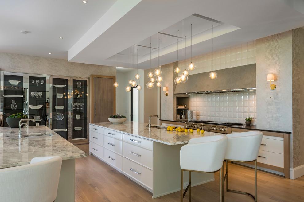 Inspiration for an expansive contemporary l-shaped open plan kitchen in Indianapolis with an undermount sink, flat-panel cabinets, white cabinets, marble benchtops, white splashback, porcelain splashback, stainless steel appliances, medium hardwood floors, multiple islands and brown floor.
