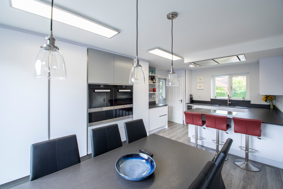 Mid-sized contemporary u-shaped eat-in kitchen in Hampshire with a drop-in sink, flat-panel cabinets, white cabinets, solid surface benchtops, black splashback, porcelain splashback, panelled appliances, light hardwood floors, a peninsula, beige floor and black benchtop.