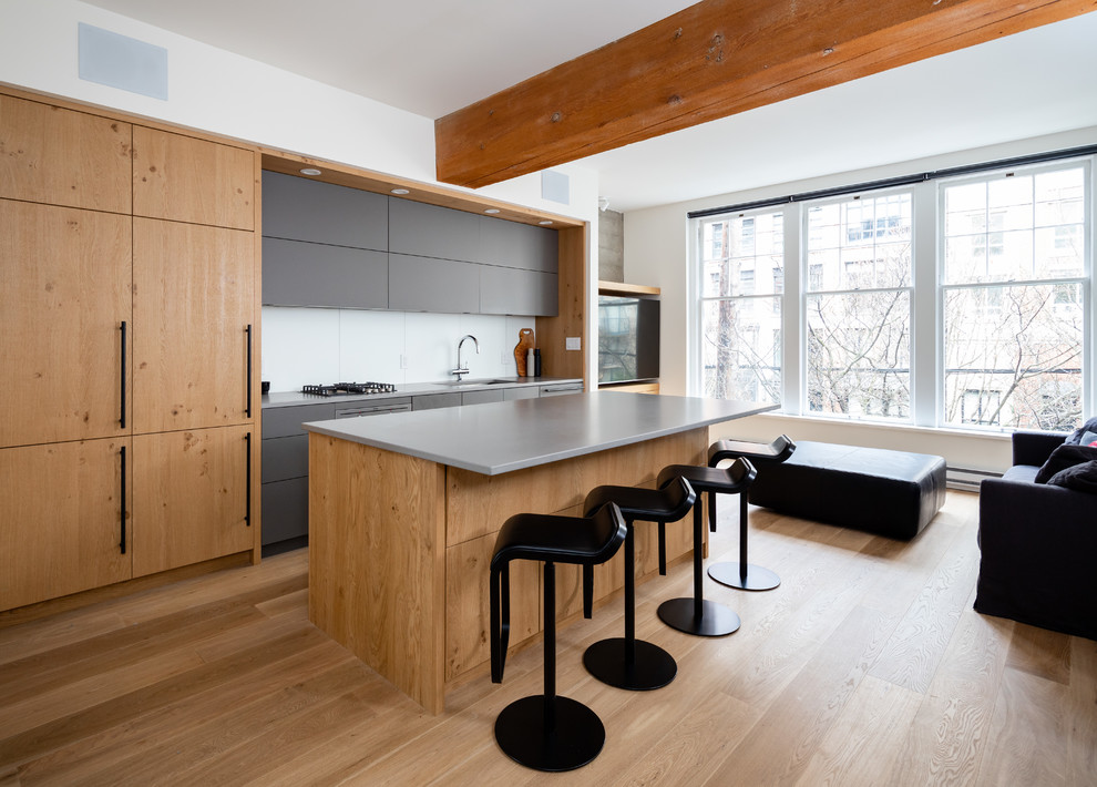 Mid-sized industrial single-wall open plan kitchen in Vancouver with flat-panel cabinets, with island, brown floor, grey benchtop, a single-bowl sink, brown cabinets, solid surface benchtops, white splashback, porcelain splashback, panelled appliances and light hardwood floors.