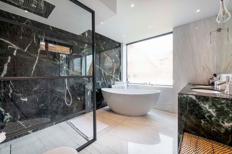 Large contemporary ensuite bathroom in London with glass-front cabinets, green cabinets, a freestanding bath, a walk-in shower, a wall mounted toilet, marble tiles, white walls, marble flooring, an integrated sink, marble worktops, white floors, an open shower, green worktops, a feature wall, double sinks and a built in vanity unit.