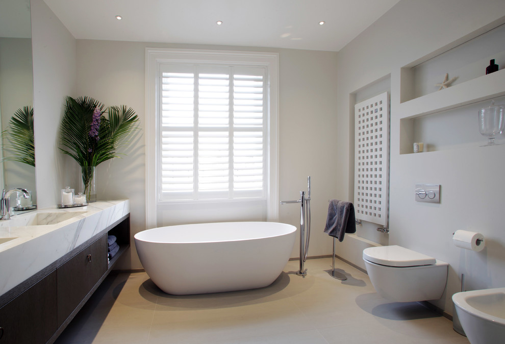 Photo of a contemporary master bathroom in London with an integrated sink, flat-panel cabinets, dark wood cabinets, a freestanding tub, a wall-mount toilet and grey walls.