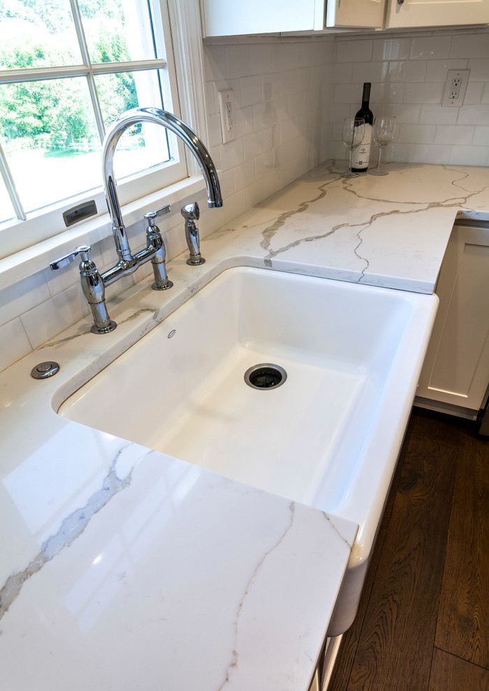 Transitional medium tone wood floor and brown floor home design photo with a farmhouse sink, white cabinets, quartz countertops, white backsplash, ceramic backsplash, stainless steel appliances and an island