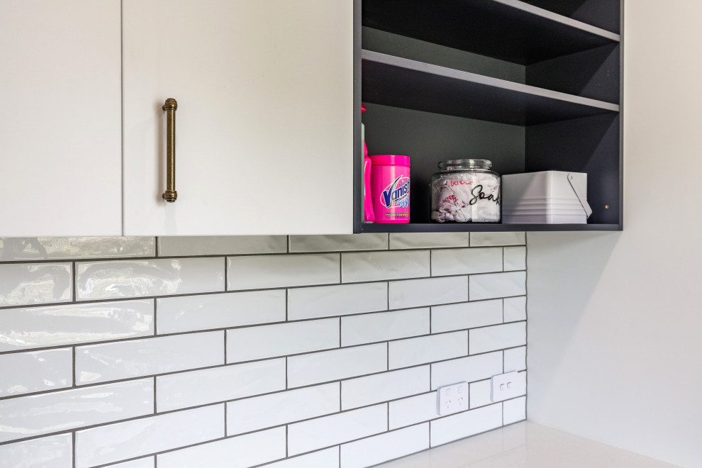 This is an example of a small traditional single-wall dedicated laundry room in Auckland with a single-bowl sink, open cabinets, white cabinets, solid surface benchtops, white splashback, subway tile splashback, white walls, porcelain floors, a side-by-side washer and dryer and white benchtop.