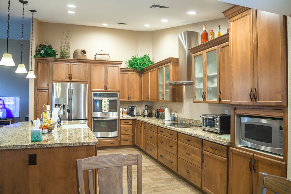 Example of a mid-sized transitional l-shaped laminate floor and brown floor open concept kitchen design in Phoenix with a farmhouse sink, recessed-panel cabinets, medium tone wood cabinets, granite countertops, beige backsplash, stone slab backsplash, stainless steel appliances and an island
