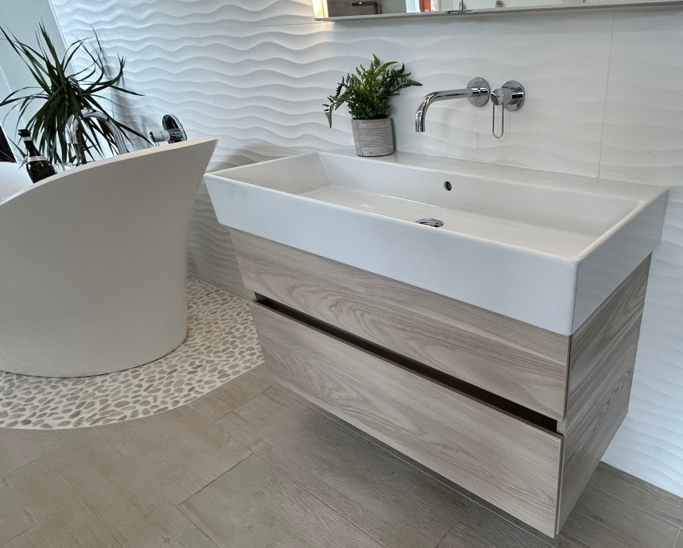 Exempel på ett stort en-suite badrum, med släta luckor, skåp i ljust trä, ett fristående badkar, vit kakel, porslinskakel, vita väggar, klinkergolv i porslin, ett avlångt handfat och beiget golv