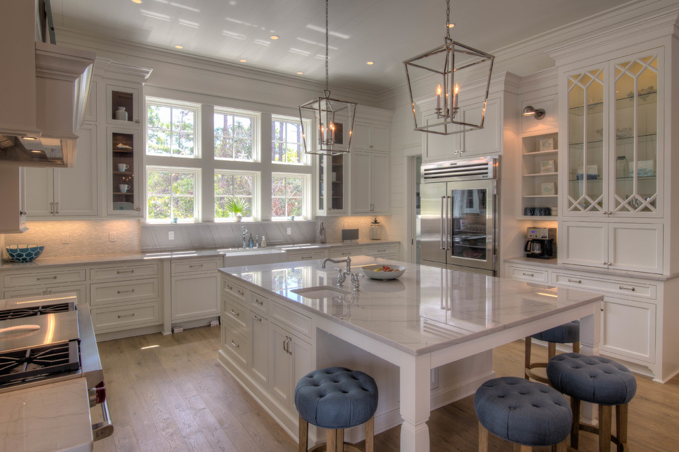 Expansive beach style u-shaped open plan kitchen in Atlanta with a farmhouse sink, recessed-panel cabinets, white cabinets, grey splashback, glass tile splashback, stainless steel appliances, light hardwood floors, with island, quartzite benchtops and beige floor.
