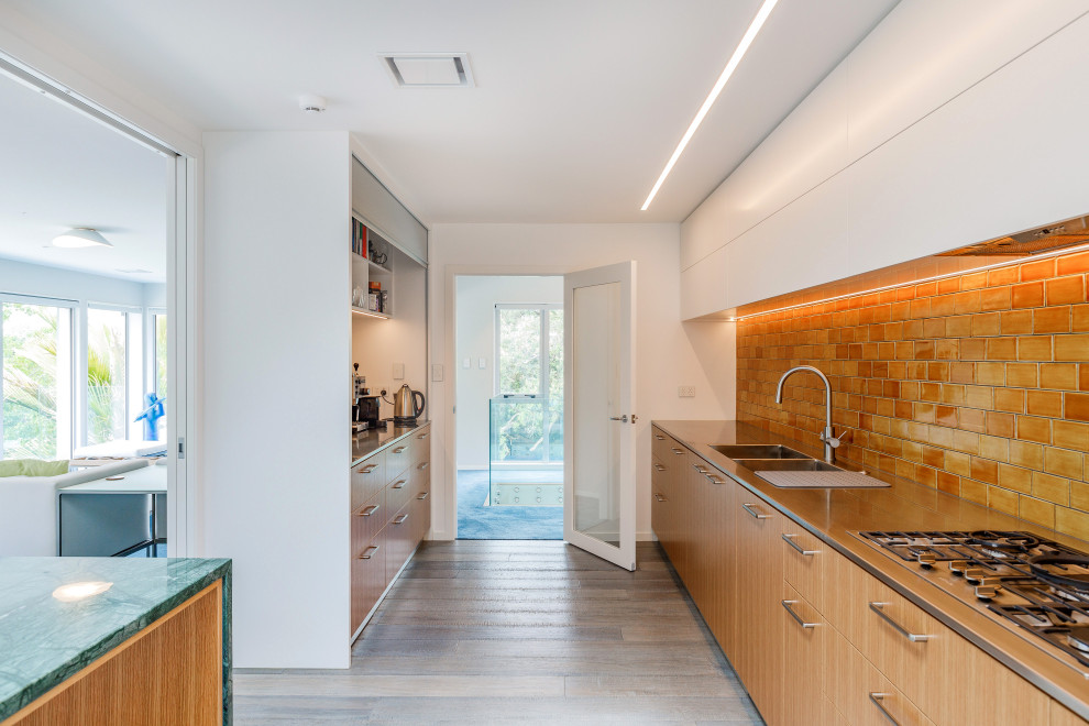Large contemporary galley kitchen pantry in Auckland with a double-bowl sink, flat-panel cabinets, medium wood cabinets, stainless steel benchtops, brown splashback, ceramic splashback, light hardwood floors, with island, blue floor and grey benchtop.