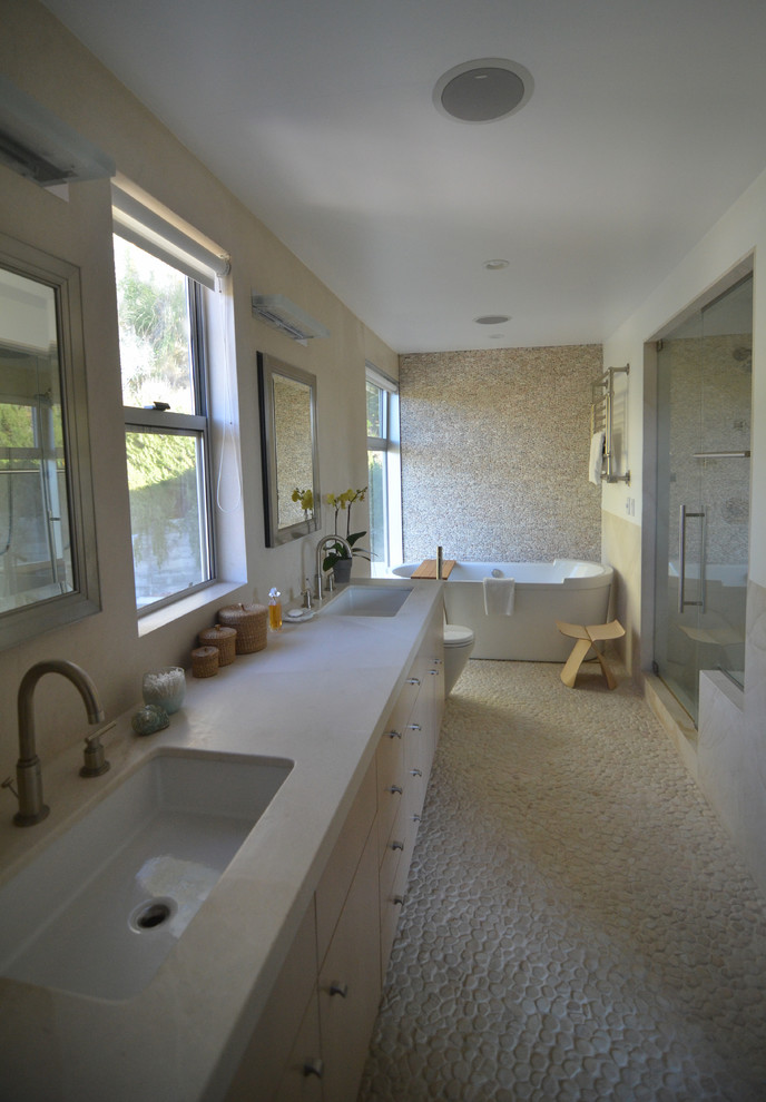 Photo of a mid-sized mediterranean bathroom in Los Angeles with flat-panel cabinets, light wood cabinets, a freestanding tub, an open shower, a two-piece toilet, beige tile, limestone, white walls, pebble tile floors, an undermount sink, limestone benchtops, beige floor and a hinged shower door.