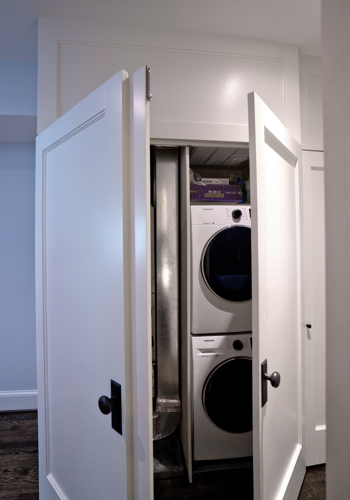 Photo of a small contemporary single-wall laundry cupboard in DC Metro with shaker cabinets, white cabinets, grey walls, dark hardwood floors, a stacked washer and dryer and brown floor.