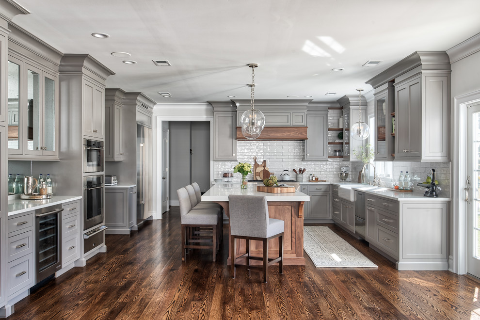 Photo of a transitional u-shaped kitchen in New York with a farmhouse sink, shaker cabinets, grey cabinets, white splashback, subway tile splashback, stainless steel appliances, medium hardwood floors, with island, brown floor and white benchtop.