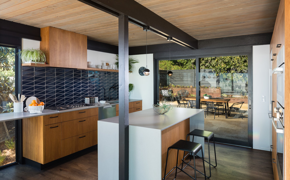 Inspiration for a midcentury galley kitchen in San Francisco with flat-panel cabinets, medium wood cabinets, black splashback, stainless steel appliances, dark hardwood floors, with island, brown floor and grey benchtop.