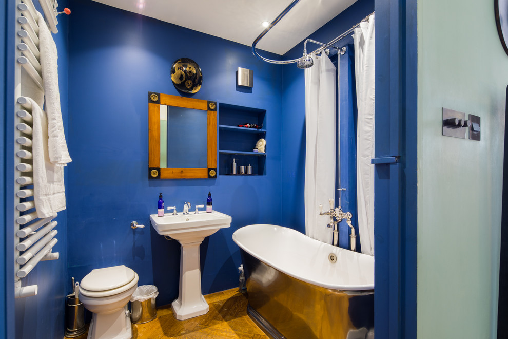 Mid-sized eclectic kids bathroom in London with open cabinets, blue cabinets, a freestanding tub, a shower/bathtub combo, a one-piece toilet, blue walls, dark hardwood floors, a pedestal sink, brown floor and a shower curtain.