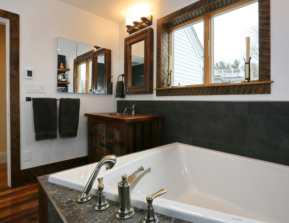 Photo of a mid-sized eclectic master wet room bathroom in Boston with furniture-like cabinets, distressed cabinets, a drop-in tub, a two-piece toilet, gray tile, stone tile, white walls, pebble tile floors, a drop-in sink and wood benchtops.