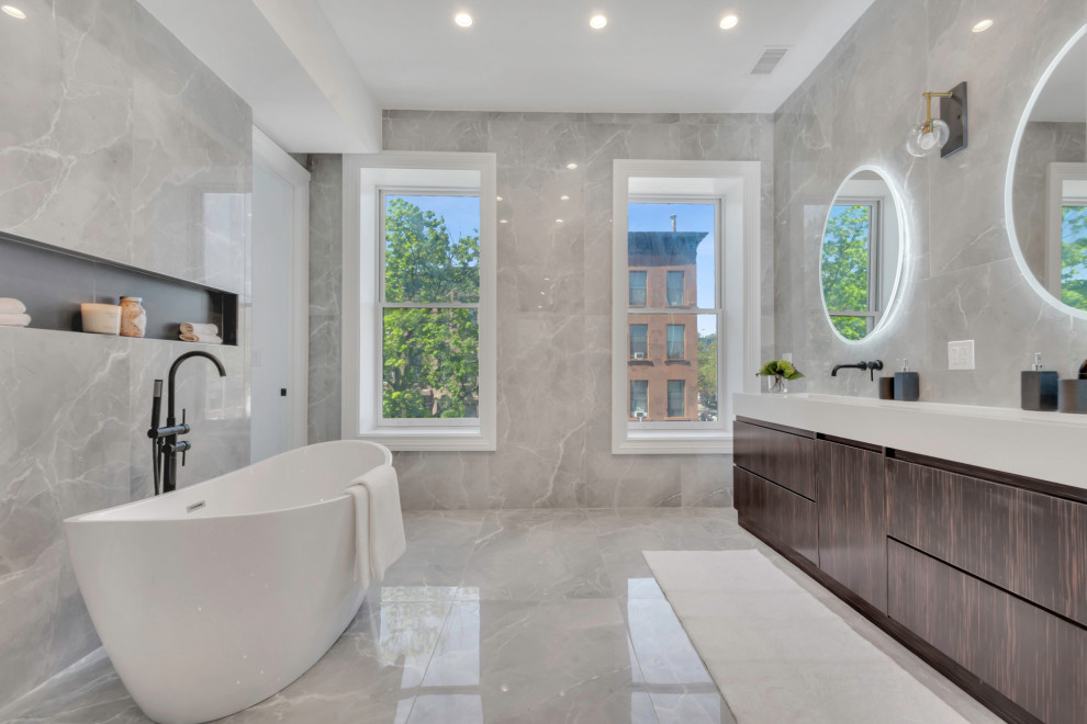 This is an example of an expansive contemporary ensuite wet room bathroom in New York with flat-panel cabinets, distressed cabinets, a freestanding bath, a wall mounted toilet, grey tiles, porcelain tiles, grey walls, porcelain flooring, a built-in sink, engineered stone worktops, grey floors, a hinged door, white worktops, double sinks and a built in vanity unit.