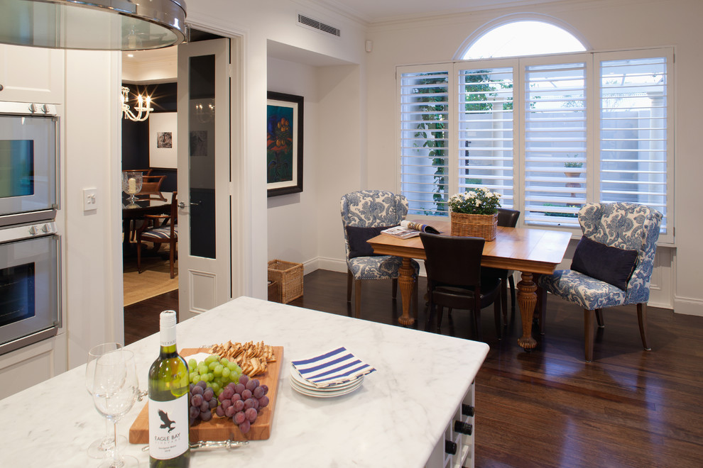 This is an example of a mid-sized country single-wall eat-in kitchen in Perth with a drop-in sink, flat-panel cabinets, white cabinets, marble benchtops, white splashback, ceramic splashback, stainless steel appliances, dark hardwood floors and with island.