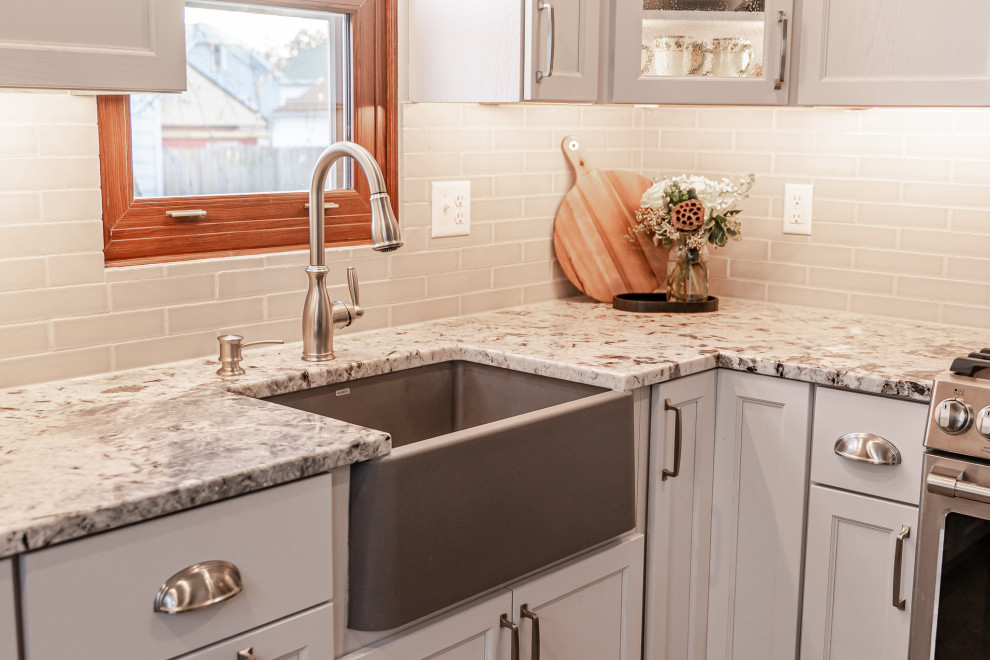 This is an example of a small country l-shaped separate kitchen in Milwaukee with a farmhouse sink, shaker cabinets, white cabinets, granite benchtops, beige splashback, ceramic splashback, stainless steel appliances, vinyl floors, no island, brown floor and multi-coloured benchtop.