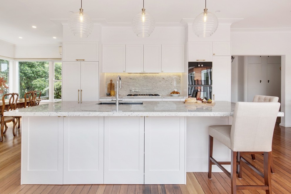 Design ideas for a mid-sized transitional single-wall open plan kitchen in Sydney with shaker cabinets, white cabinets, granite benchtops, grey splashback, marble splashback, black appliances, medium hardwood floors, with island, brown floor and white benchtop.