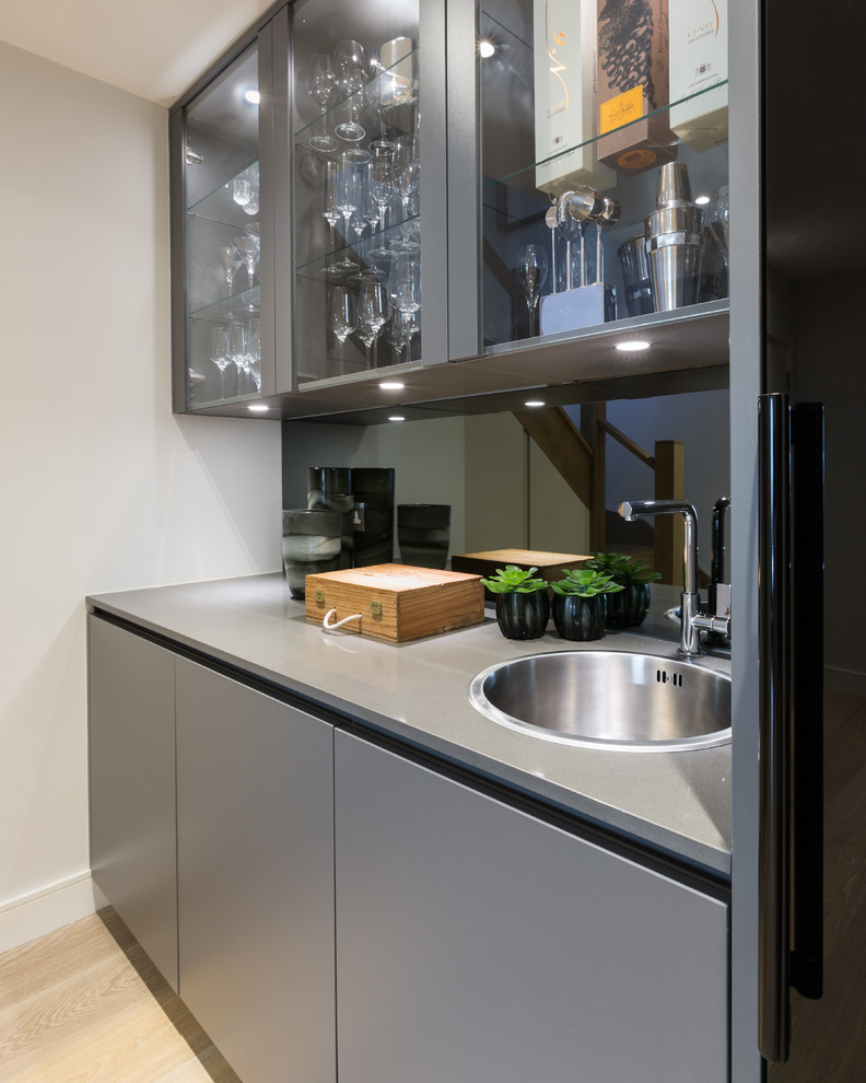 Small contemporary single-wall wet bar in Gloucestershire with a drop-in sink, flat-panel cabinets, grey cabinets, solid surface benchtops, grey splashback, mirror splashback, light hardwood floors and grey benchtop.