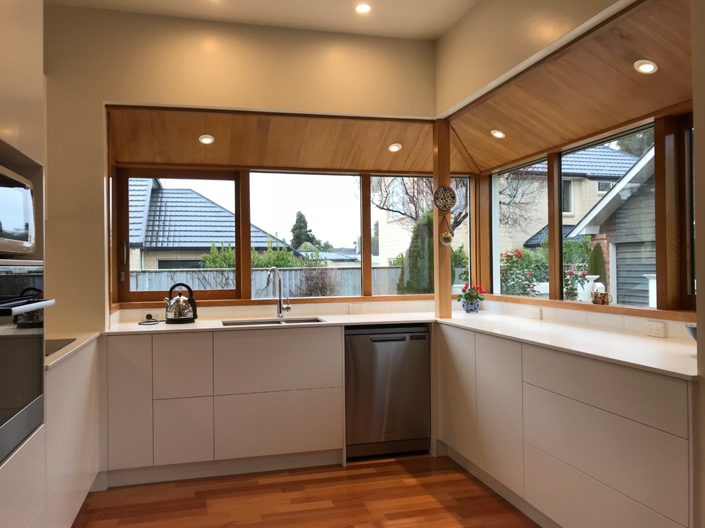 Photo of a large contemporary u-shaped open plan kitchen in Christchurch with a double-bowl sink, flat-panel cabinets, white cabinets, quartz benchtops, white splashback, glass sheet splashback, stainless steel appliances, medium hardwood floors, brown floor and white benchtop.