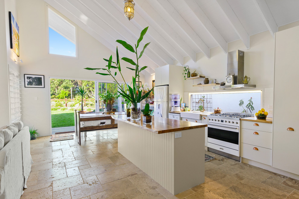 This is an example of a beach style galley kitchen in Gold Coast - Tweed with a farmhouse sink, flat-panel cabinets, white cabinets, wood benchtops, white appliances, with island, beige floor and brown benchtop.