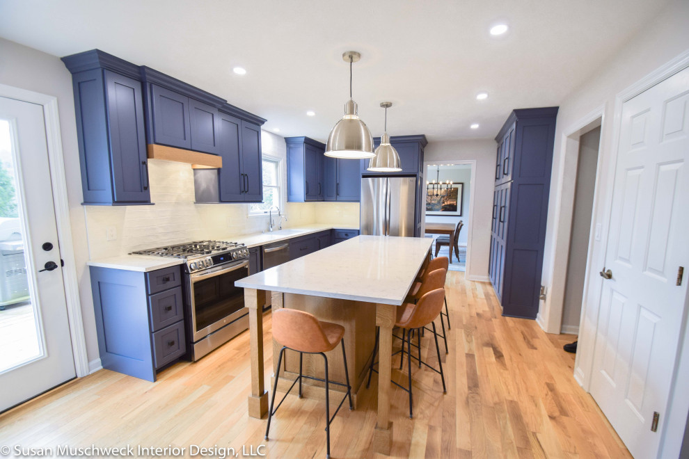 Mid-sized transitional l-shaped light wood floor enclosed kitchen photo with an undermount sink, shaker cabinets, blue cabinets, quartz countertops, white backsplash, ceramic backsplash, stainless steel appliances, an island and white countertops