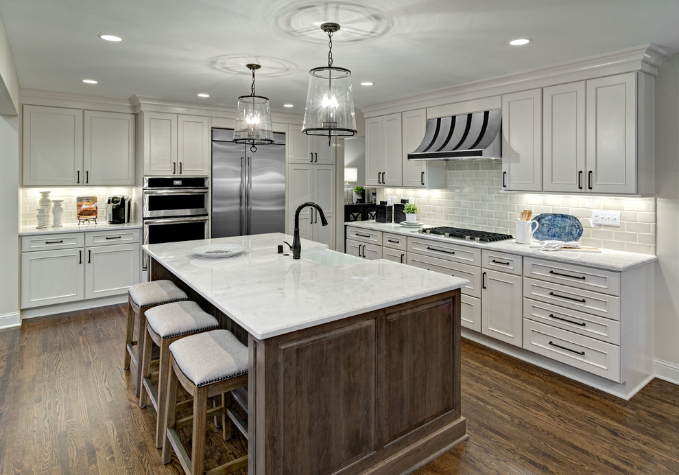 Photo of a mid-sized country l-shaped eat-in kitchen in Minneapolis with a farmhouse sink, recessed-panel cabinets, white cabinets, quartz benchtops, grey splashback, ceramic splashback, stainless steel appliances, medium hardwood floors and with island.