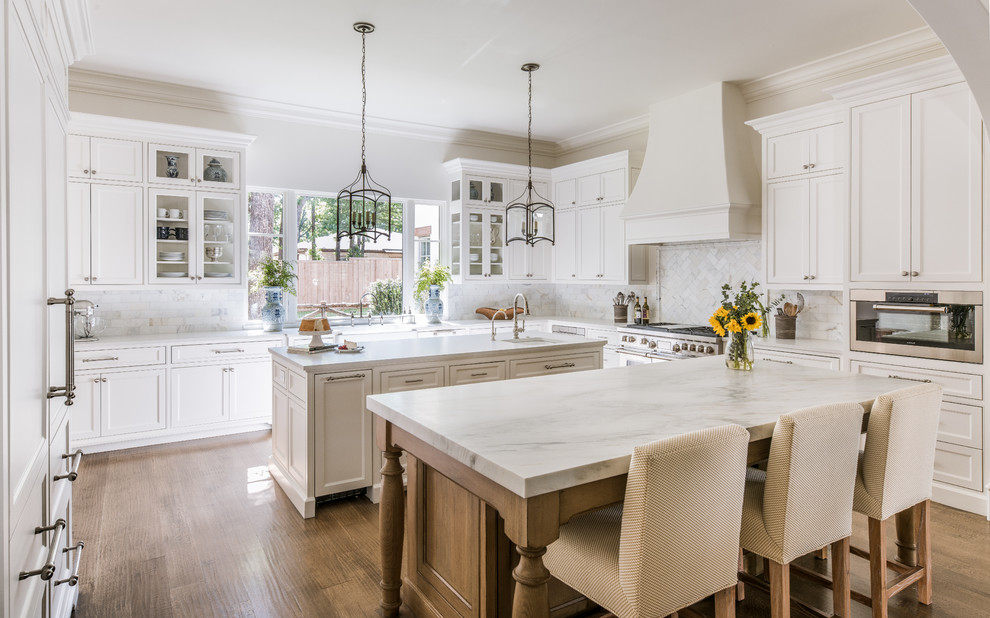 This is an example of a transitional u-shaped open plan kitchen in Dallas with a farmhouse sink, shaker cabinets, white cabinets, white splashback, panelled appliances, dark hardwood floors, multiple islands and white benchtop.