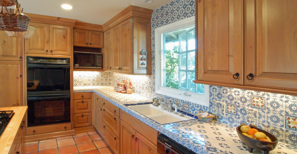 Photo of a mid-sized country l-shaped eat-in kitchen in San Luis Obispo with an undermount sink, raised-panel cabinets, light wood cabinets, tile benchtops, blue splashback, ceramic splashback, black appliances, terra-cotta floors and with island.