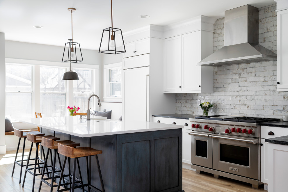 Design ideas for a large transitional galley kitchen in Denver with a farmhouse sink, shaker cabinets, white cabinets, white splashback, brick splashback, stainless steel appliances, with island, beige floor and black benchtop.