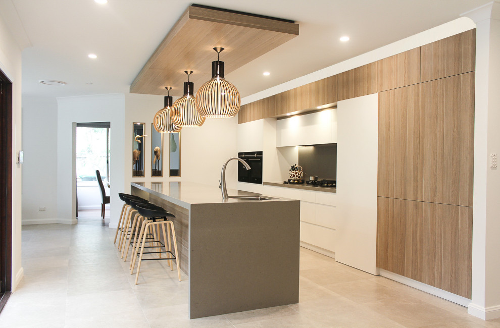 This is an example of a large contemporary single-wall open plan kitchen with an undermount sink, flat-panel cabinets, white cabinets, quartz benchtops, grey splashback, black appliances, porcelain floors, with island, grey floor and grey benchtop.