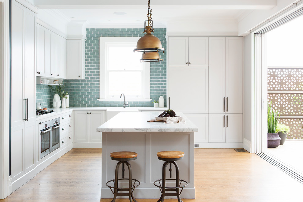 Transitional l-shaped kitchen in Sydney with a farmhouse sink, shaker cabinets, white cabinets, blue splashback, subway tile splashback, panelled appliances, medium hardwood floors, with island and brown floor.