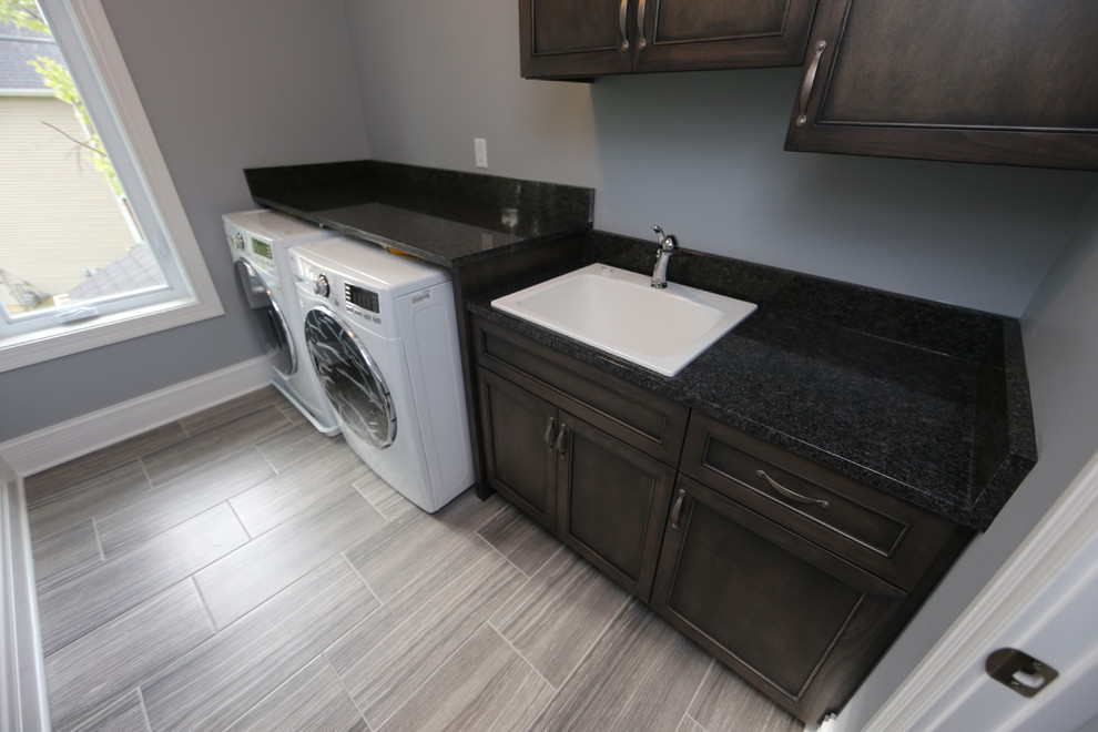 Mid-sized arts and crafts single-wall dedicated laundry room in Chicago with a drop-in sink, granite benchtops, grey walls, porcelain floors, a side-by-side washer and dryer, grey floor, black benchtop, recessed-panel cabinets and dark wood cabinets.