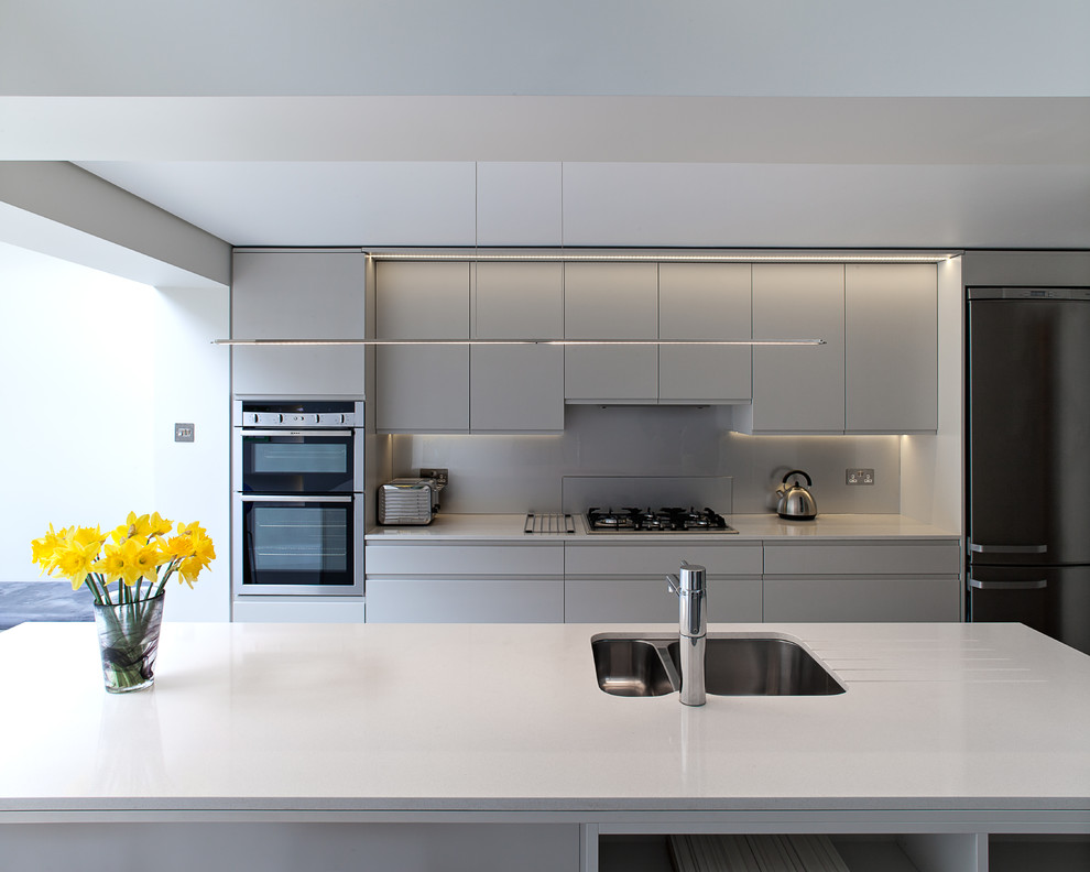 Design ideas for a large contemporary galley open plan kitchen in London with an integrated sink, flat-panel cabinets, white cabinets, quartz benchtops, grey splashback, glass sheet splashback, panelled appliances, ceramic floors, with island, beige floor and white benchtop.