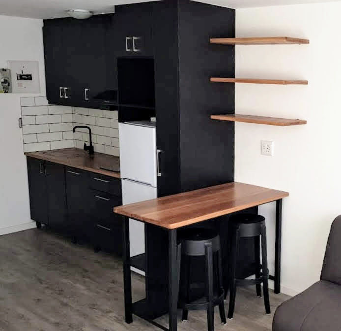 Small contemporary single-wall open plan kitchen in Other with an undermount sink, louvered cabinets, black cabinets, wood benchtops, white splashback, ceramic splashback, black appliances, vinyl floors, with island, grey floor, black benchtop and coffered.