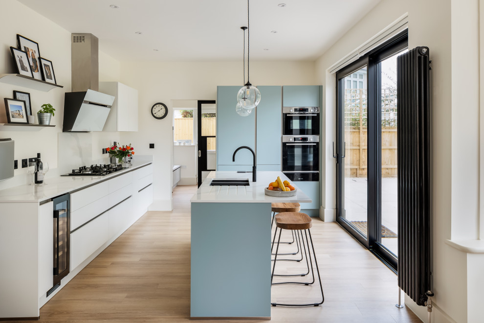 Contemporary l-shaped kitchen in Dorset with a submerged sink, flat-panel cabinets, blue cabinets, stainless steel appliances, light hardwood flooring, an island, beige floors and white worktops.