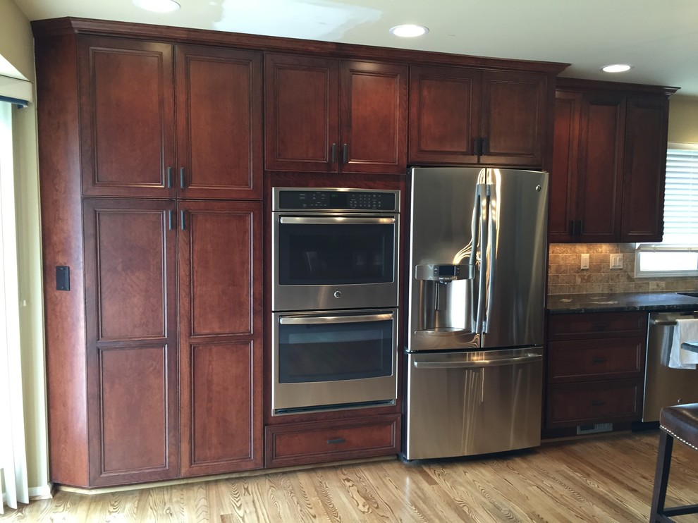This is an example of a large traditional l-shaped eat-in kitchen in Detroit with a double-bowl sink, recessed-panel cabinets, dark wood cabinets, granite benchtops, beige splashback, stone tile splashback, stainless steel appliances, light hardwood floors and with island.
