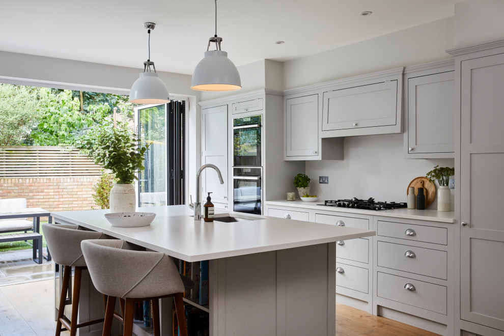 This is an example of a large transitional galley kitchen in London with shaker cabinets, grey cabinets, solid surface benchtops, white splashback, medium hardwood floors, with island, brown floor, white benchtop, an undermount sink and stainless steel appliances.