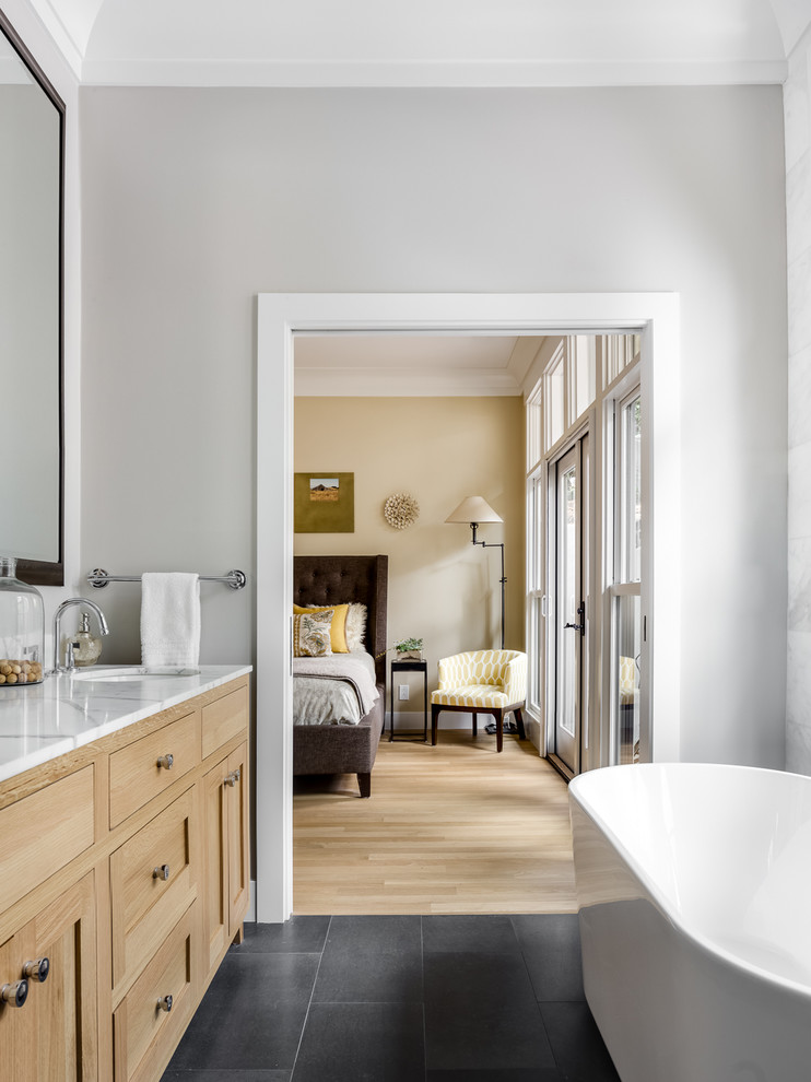 Photo of a mid-sized transitional master bathroom in San Francisco with shaker cabinets, light wood cabinets, a freestanding tub, gray tile, stone tile, grey walls, a corner shower, a one-piece toilet, limestone floors, an undermount sink and white benchtops.