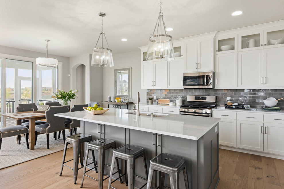 Transitional l-shaped open plan kitchen in Minneapolis with a farmhouse sink, shaker cabinets, white cabinets, quartzite benchtops, grey splashback, subway tile splashback, stainless steel appliances, with island, white benchtop, medium hardwood floors and brown floor.