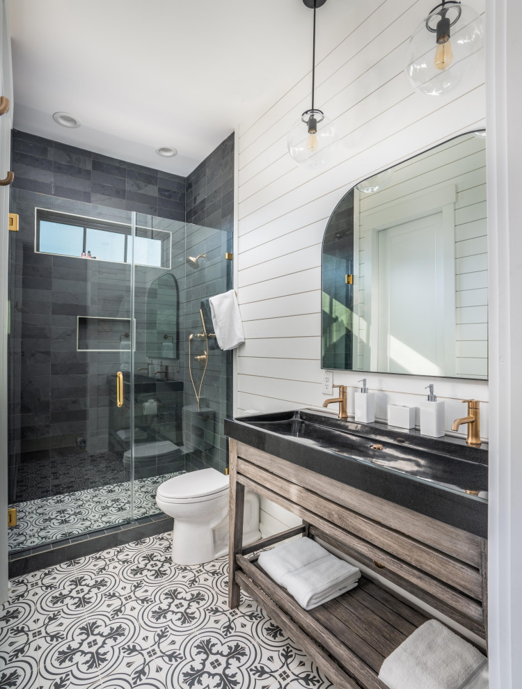 Photo of a large transitional 3/4 bathroom in Charleston with open cabinets, medium wood cabinets, an alcove shower, a two-piece toilet, gray tile, white walls, a trough sink, multi-coloured floor, a hinged shower door, black benchtops, a niche, a single vanity, a built-in vanity and planked wall panelling.